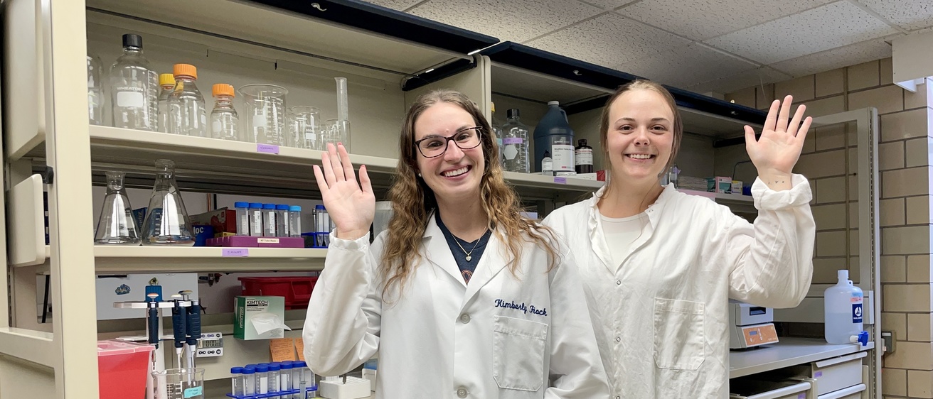 Kim and Mikayla, two lab members in lab coats, waving
