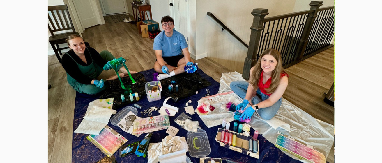 Three people smiling while sitting on a tarp with craft supplies in a house.