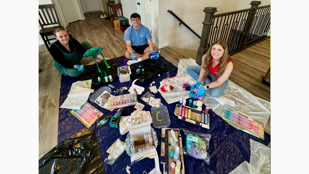 Three people smiling while sitting on a tarp with craft supplies in a house.
